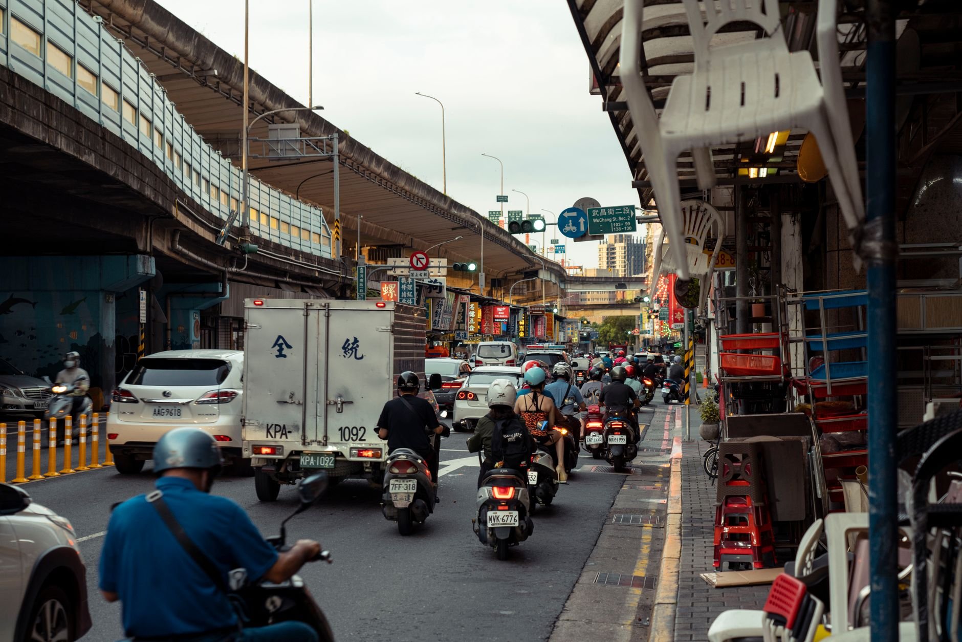 A bustling city street filled with motor scooters and vehicles under an overpass, capturing urban life.
